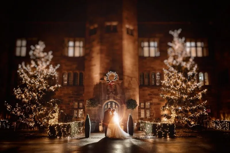 Bride and groom embracing by the Christmas trees and stone archway during a winter wedding at Abbey House Hotel